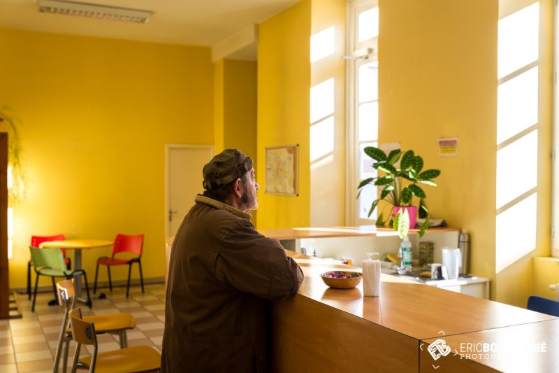 Un usager accoudé au comptoir de la salle commune, dans la lumière des fenêtres.