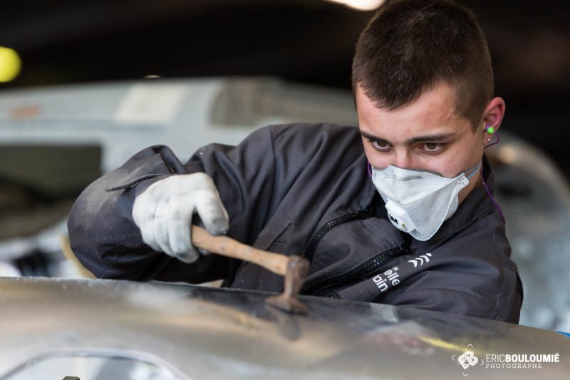 Un apprenti carrossier débosselle un panneau au marteau, masque et gants de protection.