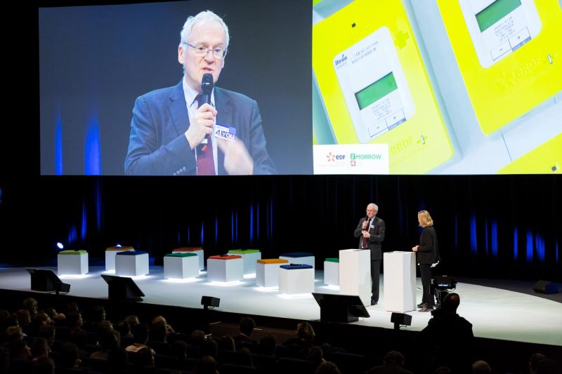 Intervenant au micro sur la scène d’un auditorium, son image projetée sur un écran géant face au public