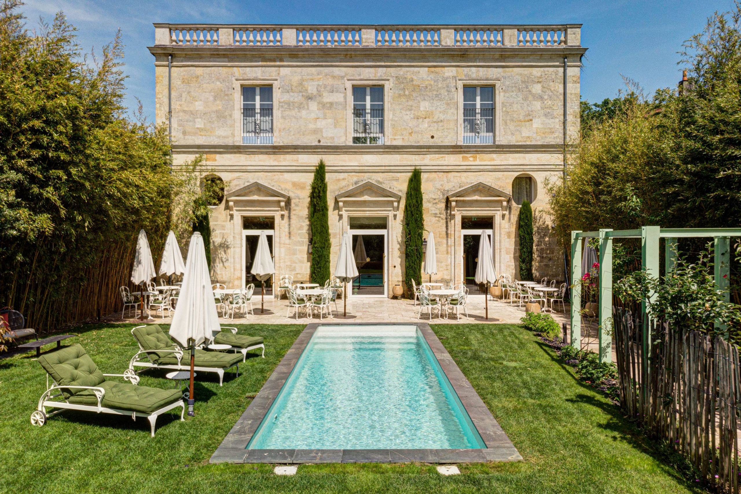 Maison de maître en pierre vue en surplomb du jardin, piscine, transats et cyprès devant la façade