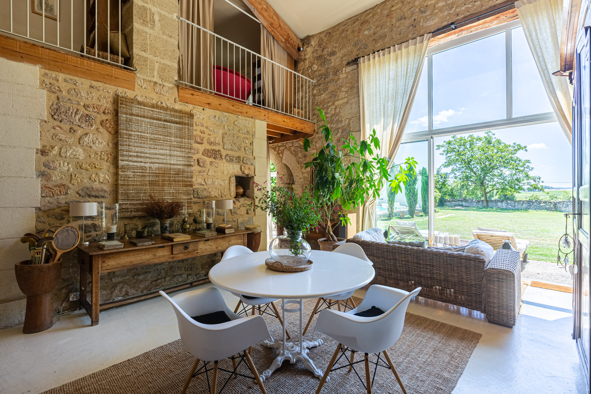 Séjour sous haut plafond : mur de pierre, mezzanine à garde-corps, grande baie sur le jardin