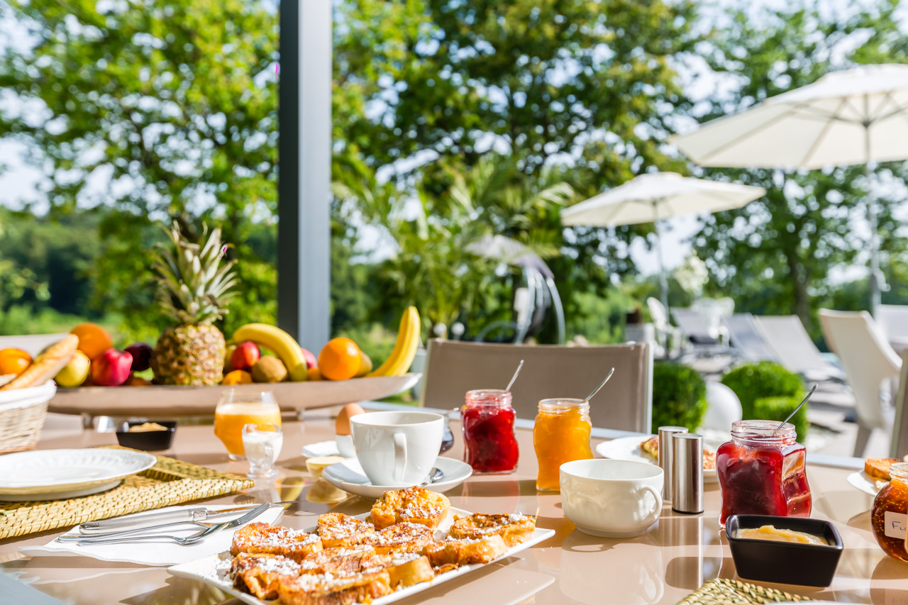 Petit-déjeuner servi devant une baie vitrée : fruits, confitures et pain perdu, terrasse et parasols au fond
