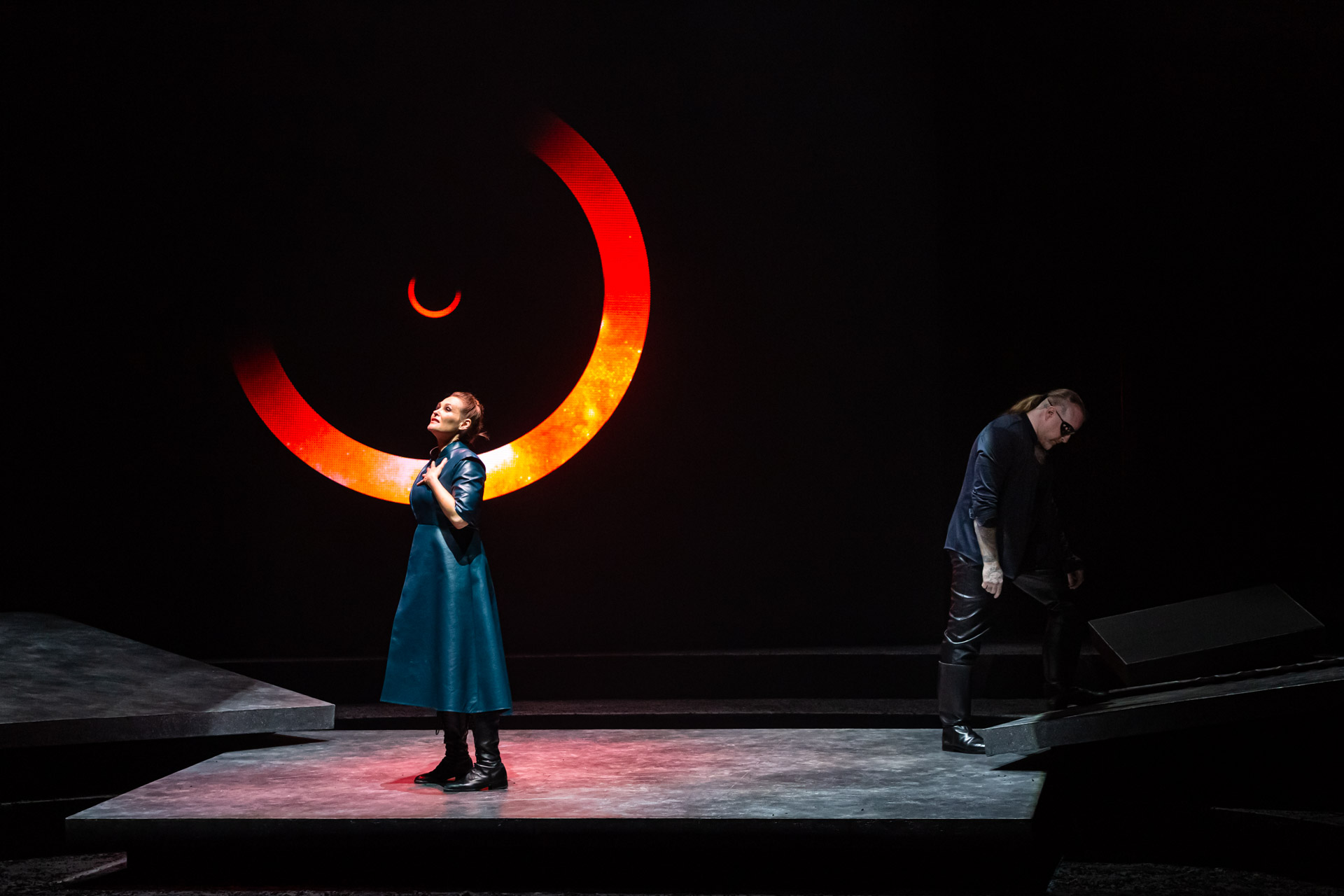 Une interprète en manteau bleu sous un grand anneau lumineux orange, un homme à l’écart sur le plateau
