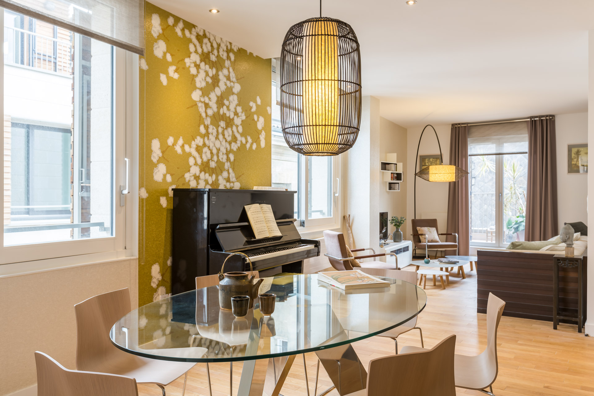 Séjour traversant : table ovale en verre, piano droit contre un mur jaune à motif floral, suspension en cage