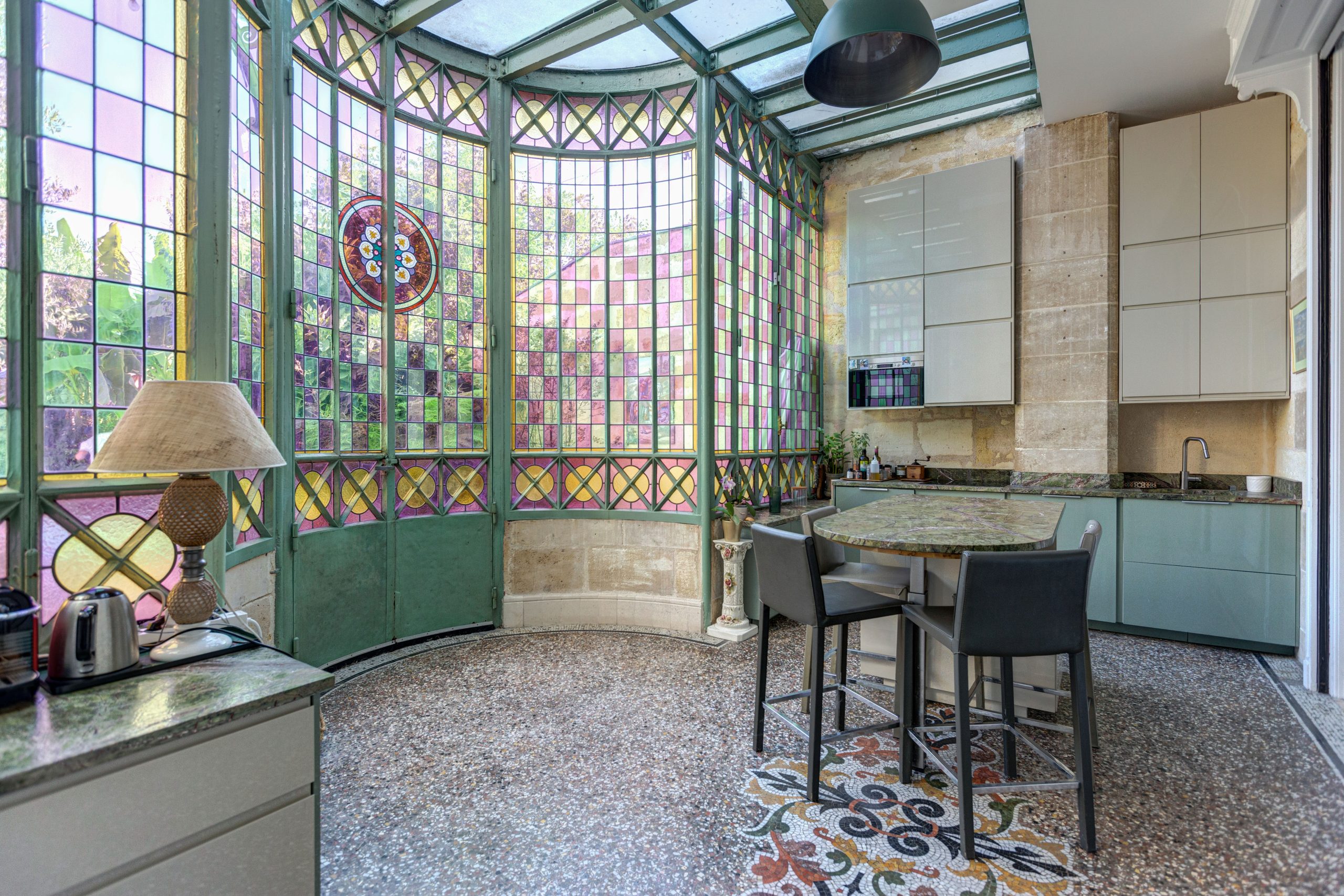 Cuisine dans une véranda à vitraux colorés, sol en mosaïque, îlot et tabourets
