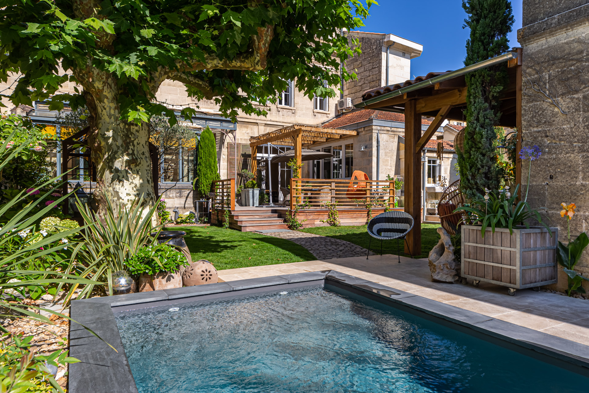 Jardin d’une bâtisse en pierre : petite piscine, pergolas et terrasse sous un platane