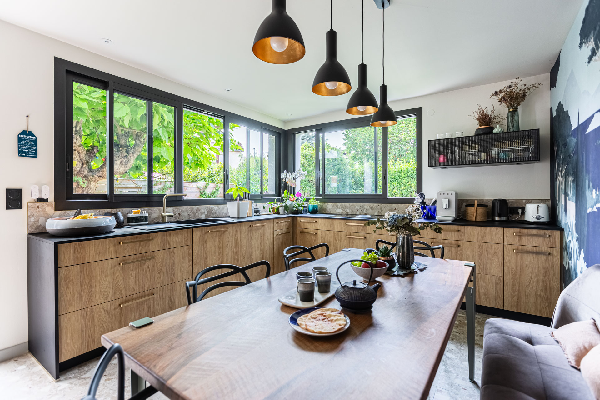 Cuisine en bois, suspensions et grande table devant des fenêtres d’angle