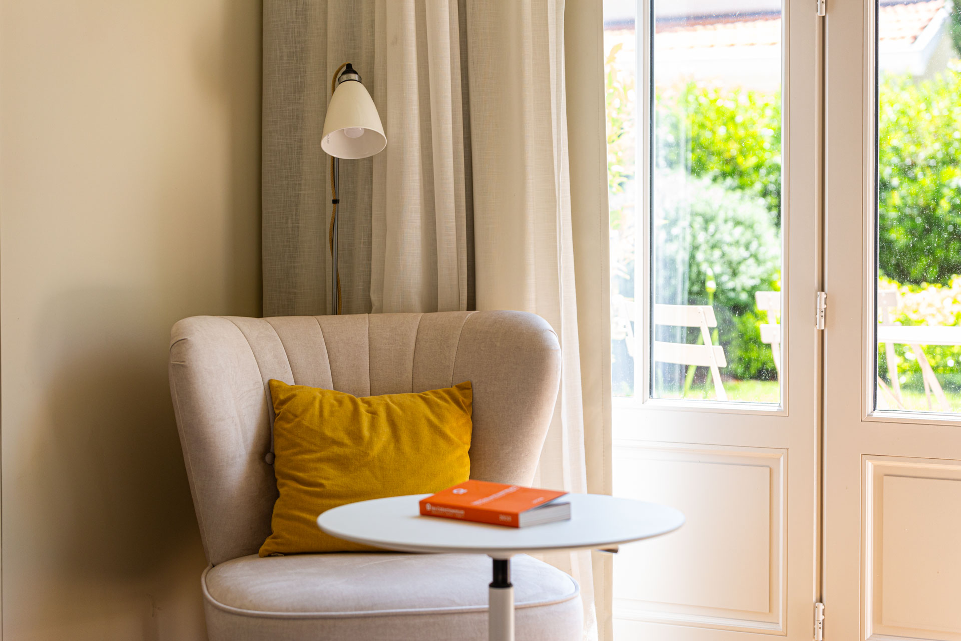 Coin lecture : fauteuil crème et coussin jaune près d’une porte-fenêtre ouverte sur le jardin