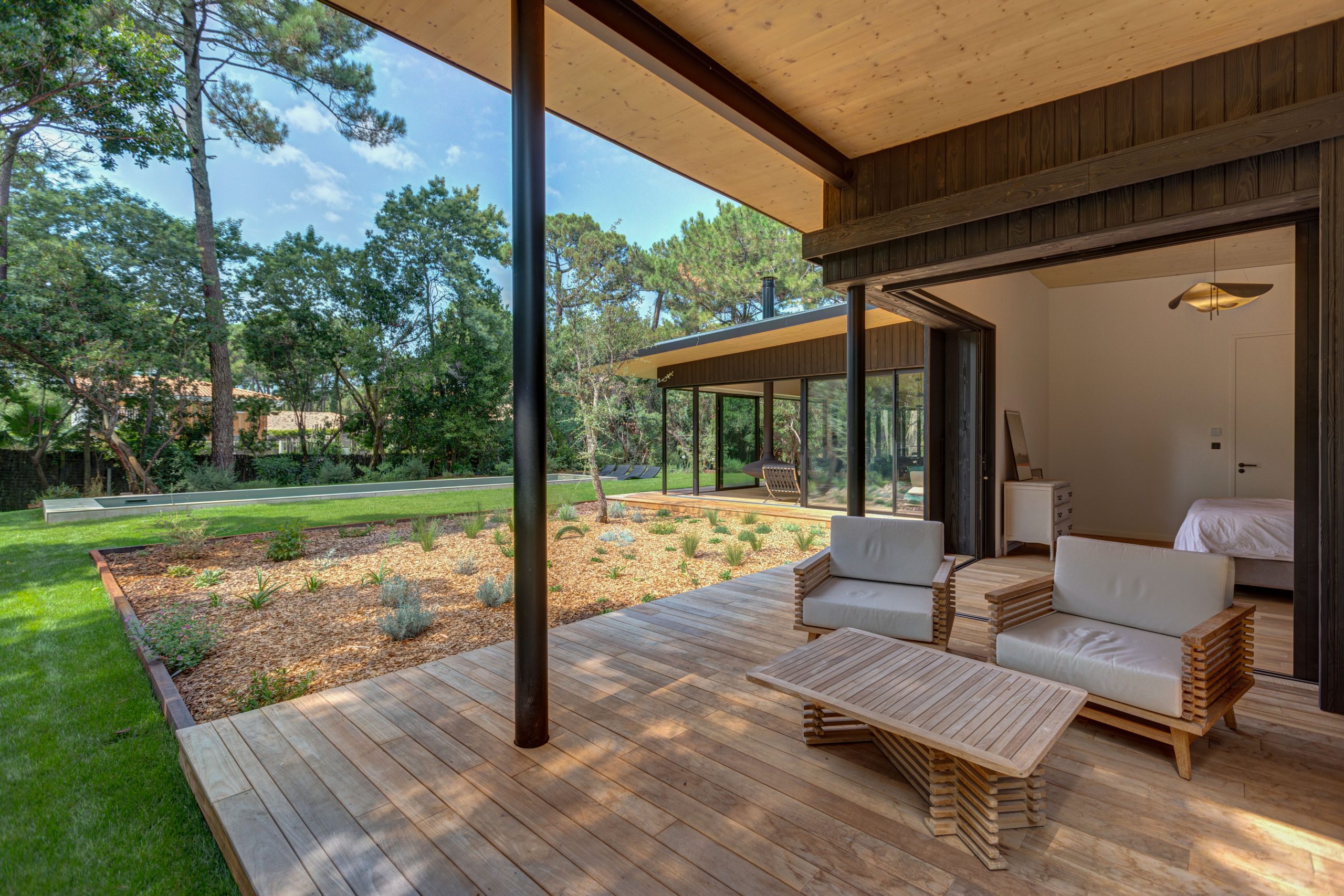 Terrasse couverte sous une charpente de bois, fauteuils, jardin et pins