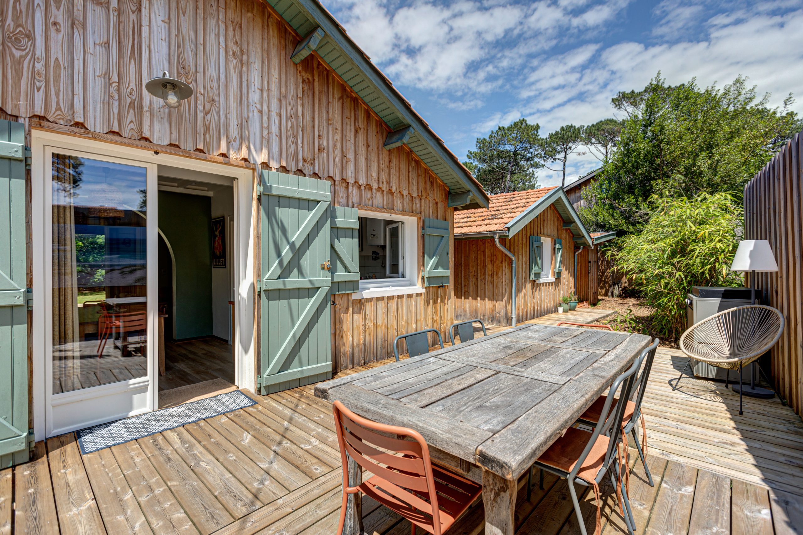 Maison à bardage bois aux volets verts, terrasse en lames, grande table et chaises