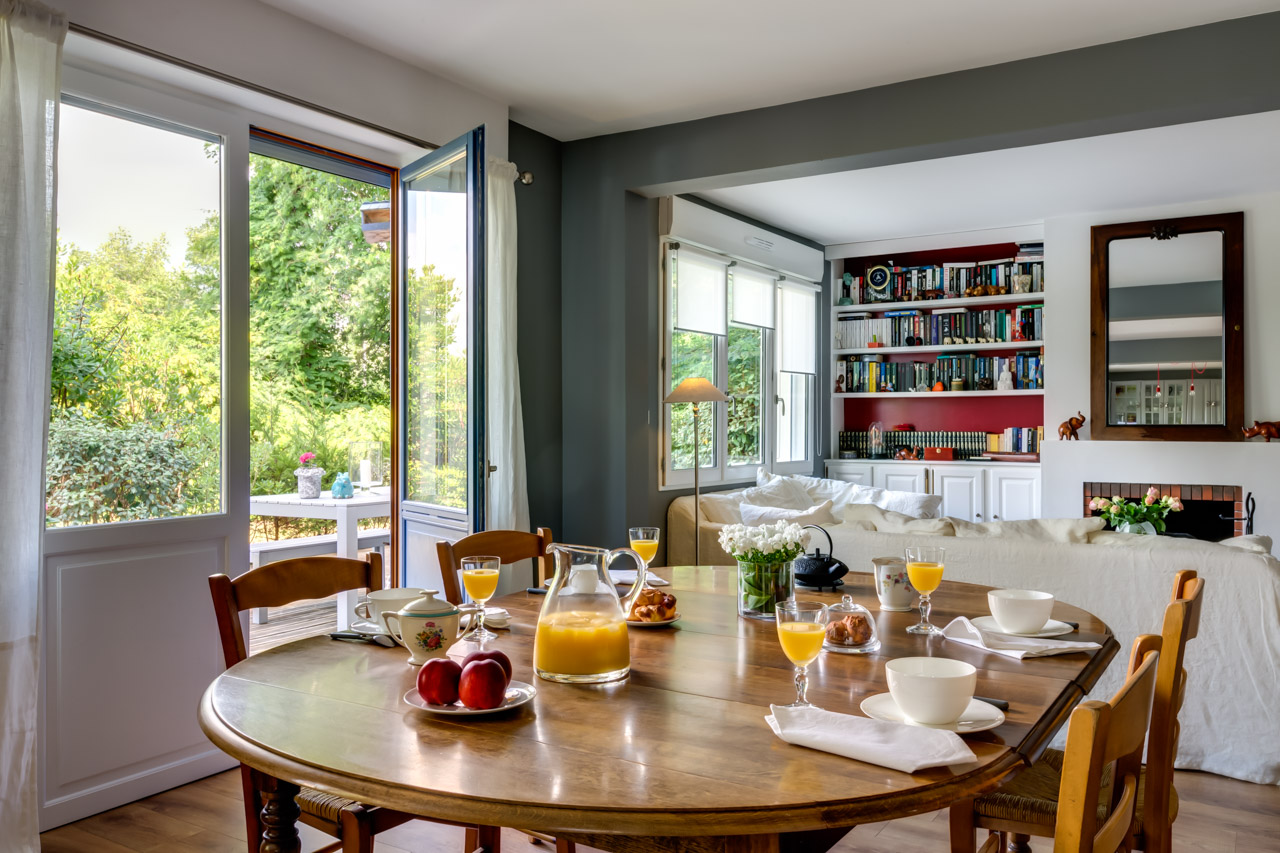 Séjour ouvert sur le jardin par des portes-fenêtres, table ronde dressée pour le petit-déjeuner, bibliothèque au fond