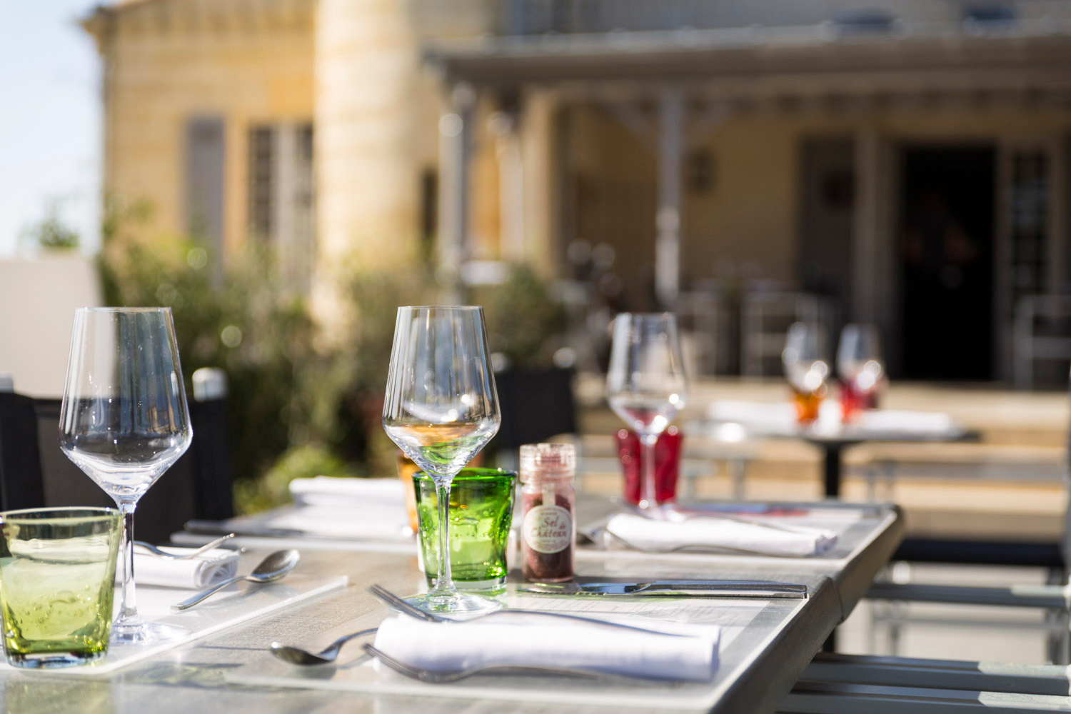 Table dressée en terrasse, verres à vin et serviettes blanches, façade en pierre au fond