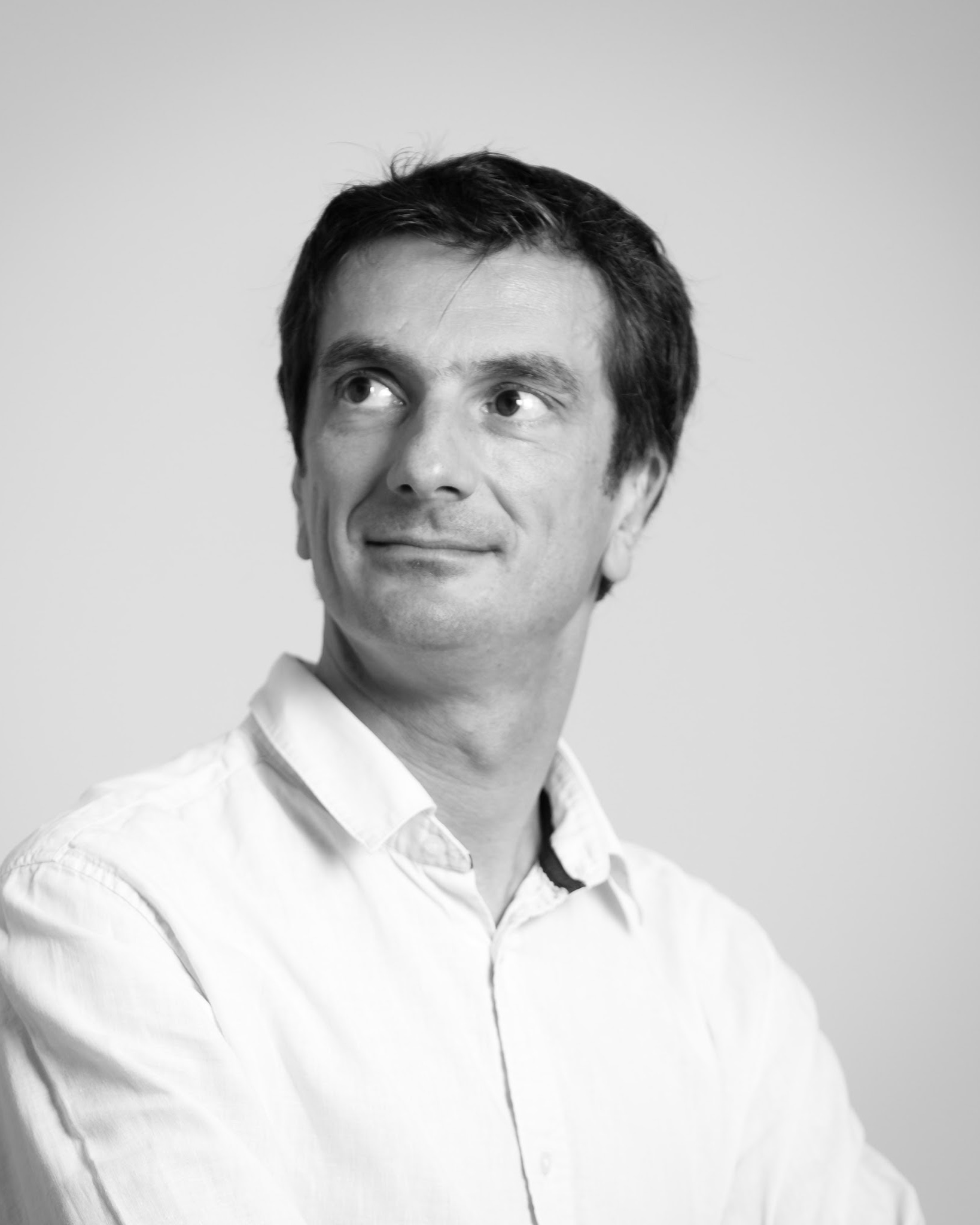 Portrait en noir et blanc d’Eric Bouloumié en chemise blanche, le regard tourné vers le haut à droite, sur fond gris clair